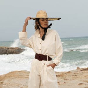 Camisa Miyako beige hecha algodon organico con mangas corte japones, puño vuelto y cuello con cuerpo para mantenerse alto, modelo con outfit negro y sombrero en la playa
